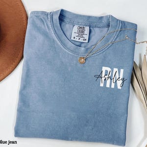 May include: A blue jean t-shirt with the text "Ashley RN" in white lettering. The shirt has a distressed look and is made of a soft, comfortable fabric.