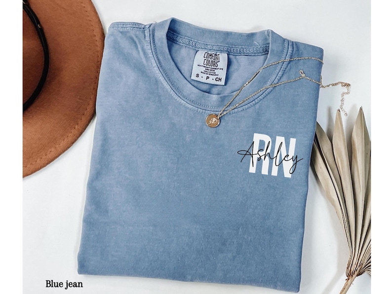 May include: A blue jean t-shirt with the text "Ashley RN" in white lettering. The shirt has a distressed look and is made of a soft, comfortable fabric.