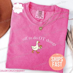 Op de afbeelding: Een felroze T-shirt met korte mouwen en een afbeelding van een witte gans die een koffiebeker vasthoudt. De tekst boven de gans luidt off to do OT things in witte letters. Het T-shirt heeft een ronde hals en een Comfort Colors label.