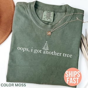 Könnte beinhalten: Ein moosgrünes T-Shirt mit dem Text "oops, i got another tree" und einer Weihnachtsbaumgrafik. Das Shirt hat einen Rundhalsausschnitt und kurze Ärmel. Eine goldene Halskette mit Anhänger liegt auf dem Shirt.