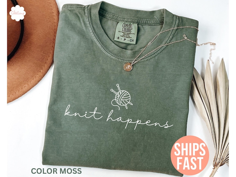 May include: A moss green t-shirt with the words "knit happens" in white script. The shirt features a graphic of yarn and knitting needles. A gold necklace with a pendant rests on the shirt. A brown hat is visible in the background.