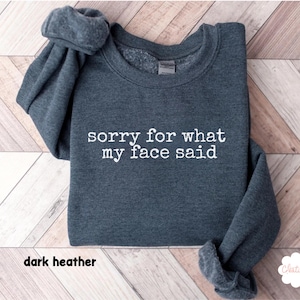 May include: Dark heather gray sweatshirt with the text "sorry for what my face said" in white. The sweatshirt has a crew neck and long sleeves. The words "dark heather" are at the bottom of the image.