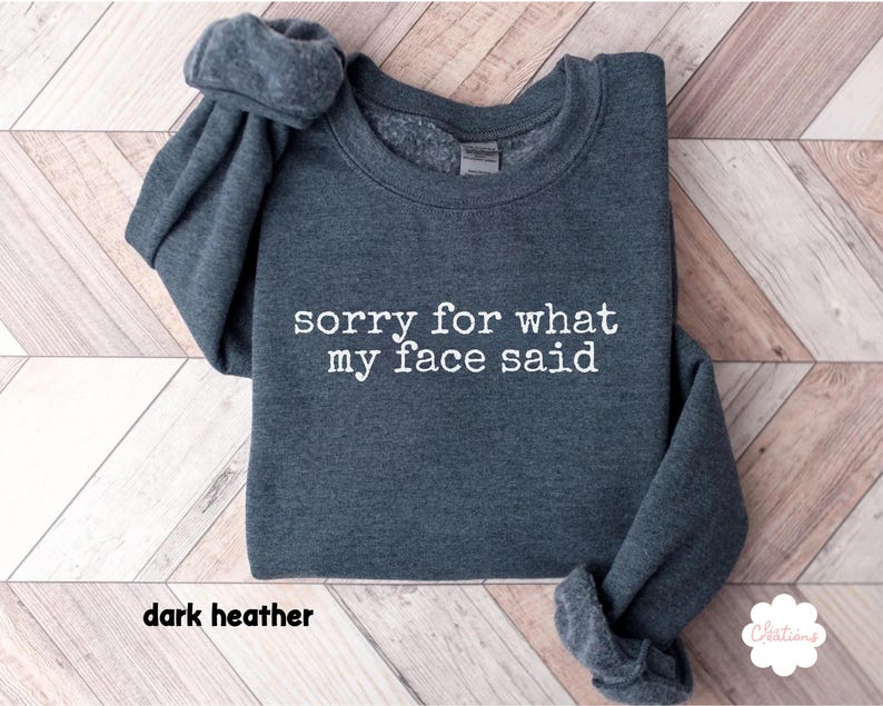 May include: Dark heather gray sweatshirt with the text "sorry for what my face said" in white. The sweatshirt has a crew neck and long sleeves. The words "dark heather" are at the bottom of the image.
