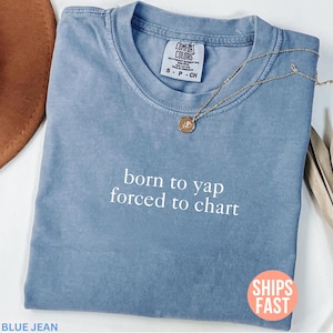 May include: A blue jean Comfort Colors t-shirt with the text "born to yap forced to chart" in white. A gold necklace with a pendant rests on the shirt. The shirt is folded, and a brown hat is partially visible.