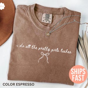Op de afbeelding: Espresso-kleurige t-shirt met de tekst "i do all the pretty girls lashes" in witte letters. Een zilveren ketting met een kleine hanger ligt op de shirt. De shirt heeft een ronde hals en korte mouwen.