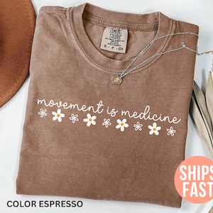 May include: Espresso-colored t-shirt with the phrase "movement is medicine" in white script and daisy graphics. A silver necklace with a pendant rests on the shirt. The shirt is a Comfort Colors brand.