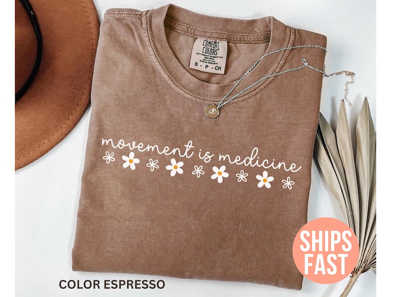 May include: Espresso-colored t-shirt with the phrase "movement is medicine" in white script and daisy graphics. A silver necklace with a pendant rests on the shirt. The shirt is a Comfort Colors brand.
