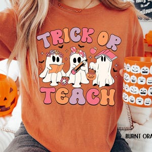 May include: Burnt orange t-shirt with the words "Trick or Teach" in a retro font. The shirt features three cartoon ghosts, one reading a book, one with a pencil, and one with a pumpkin bucket. A matching pumpkin mug is held in the hand.