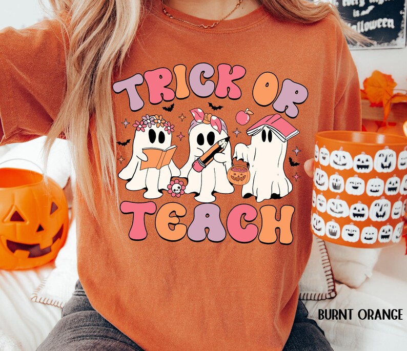 May include: Burnt orange t-shirt with the words "Trick or Teach" in a retro font. The shirt features three cartoon ghosts, one reading a book, one with a pencil, and one with a pumpkin bucket. A matching pumpkin mug is held in the hand.