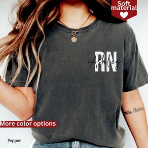 May include: A dark gray t-shirt with the text "RN" and "Ashley" in white lettering. The shirt is made of a soft material.