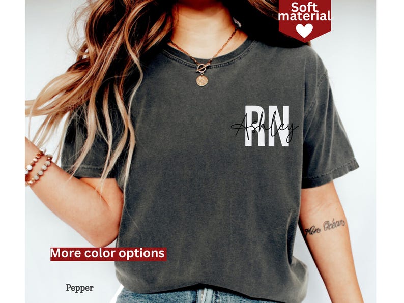 May include: A dark gray t-shirt with the text "RN" and "Ashley" in white lettering. The shirt is made of a soft material.
