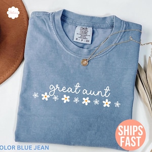 May include: A blue jean colored t-shirt with the words "great aunt" in white script, decorated with white daisy flowers. A gold necklace with a pendant rests on the shirt. The shirt is a Comfort Colors brand.