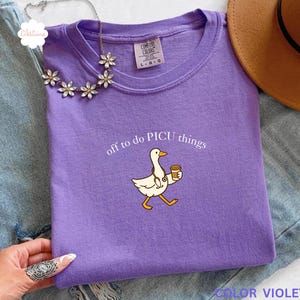 May include: A color violet t-shirt with the text "off to do PICU things" and a cartoon duck wearing a stethoscope and holding a coffee cup. The shirt is folded on a denim surface, with a necklace and a brown hat in the background.