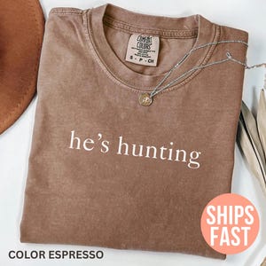 May include: Espresso-colored t-shirt with the phrase "he's hunting" in white. A silver necklace with a pendant rests on the shirt. A brown hat and dried plant stems are in the background.