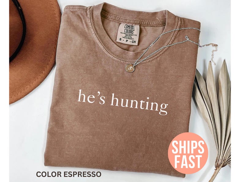 May include: Espresso-colored t-shirt with the phrase "he's hunting" in white. A silver necklace with a pendant rests on the shirt. A brown hat and dried plant stems are in the background.