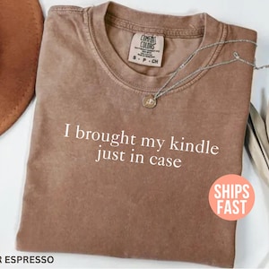 Könnte beinhalten: Braunes T-Shirt mit dem Text "I brought my kindle just in case". Das Shirt hat einen Rundhalsausschnitt und ein kleines Etikett. Eine silberne Halskette mit Anhänger befindet sich auf dem Shirt. Ein brauner Hut ist im Hintergrund zu sehen.
