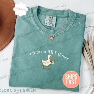 May include: A light green crew neck t-shirt featuring a graphic of a white goose holding a brown coffee cup. The text above the goose reads "off to do RBT things" in white lettering. The shirt has a Comfort Colors brand label at the neckline.