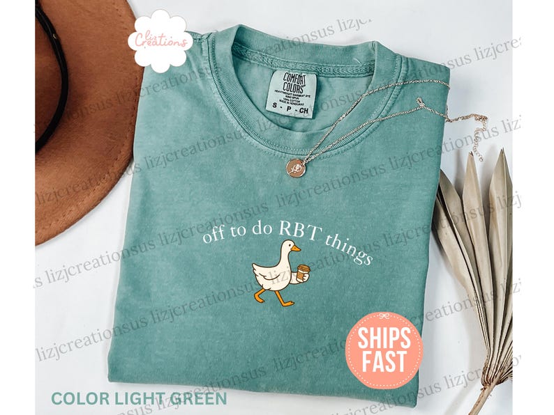 May include: A light green crew neck t-shirt featuring a graphic of a white goose holding a brown coffee cup. The text above the goose reads "off to do RBT things" in white lettering. The shirt has a Comfort Colors brand label at the neckline.