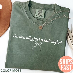 May include: An olive green t-shirt with the text "i'm literally just a hairstylist" in white and a bow graphic. The shirt is a Comfort Colors brand, size S-P-CH. A gold necklace with a pendant is on the shirt. The words "SHIPS FAST" are on a pink circle.