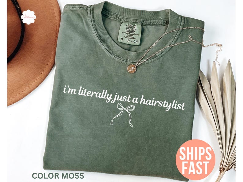 May include: An olive green t-shirt with the text "i'm literally just a hairstylist" in white and a bow graphic. The shirt is a Comfort Colors brand, size S-P-CH. A gold necklace with a pendant is on the shirt. The words "SHIPS FAST" are on a pink circle.