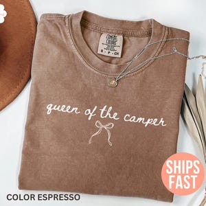 May include: Espresso-colored t-shirt with the text "queen of the camper" in white script. A silver necklace with a pendant rests on the shirt. The shirt is folded and displayed with a brown hat.