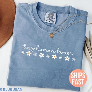 Op de afbeelding: Een blauw spijkerbroekkleurig T-shirt met de tekst "tiny human tamer" in witte letters, versierd met witte madeliefjes. Een gouden ketting met een hanger is over het shirt gedrapeerd. Het shirt is opgevouwen, met een bruine hoed en een gedroogde plant op de achtergrond.