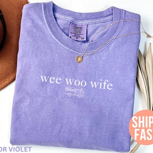 May include: A violet Comfort Colors t-shirt with the phrase "wee woo wife" and a fire truck graphic. A gold necklace with a pendant is draped on the shirt. A brown hat is visible in the background.