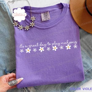 May include: A violet-colored t-shirt with the text "it's a great day to play mahjong" in white script and daisy graphics. The shirt is a crew neck and is labeled "Color Violet". A floral necklace is draped over the shirt.