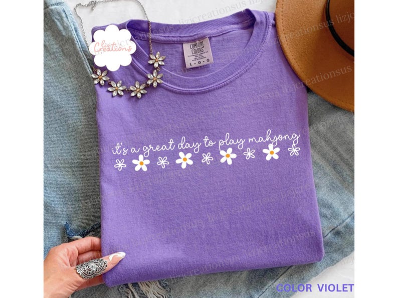 May include: A violet-colored t-shirt with the text "it's a great day to play mahjong" in white script and daisy graphics. The shirt is a crew neck and is labeled "Color Violet". A floral necklace is draped over the shirt.