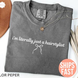 May include: Grey Comfort Colors t-shirt with the text "i'm literally just a hairstylist" in white. A silver necklace with a pendant is on the shirt. An orange sticker says "SHIPS FAST".