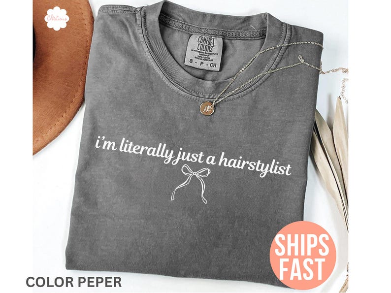 May include: Grey Comfort Colors t-shirt with the text "i'm literally just a hairstylist" in white. A silver necklace with a pendant is on the shirt. An orange sticker says "SHIPS FAST".