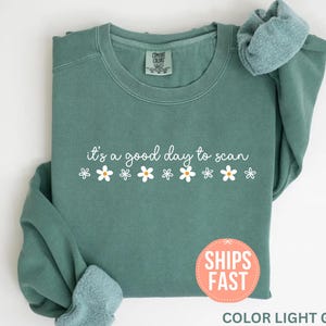 Puede incluir: Una sudadera verde claro con la frase "it's a good day to scan" en escritura blanca, decorada con gráficos de margaritas blancas. La sudadera tiene cuello redondo y mangas largas. Las palabras "Ships Fast" están en una pegatina redonda naranja.
