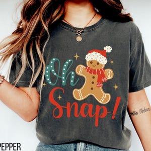 Può includere: T-shirt grigio scuro con un omino di pan di zenzero con sciarpa e cappello rossi, con le parole "Oh Snap!" in verde acqua e rosso. La maglietta ha un aspetto vintage.