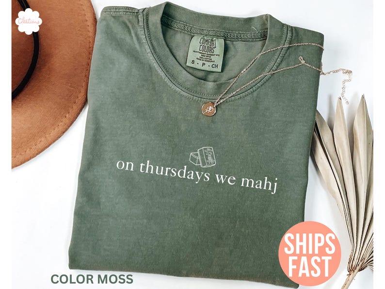 以下が含まれることがあります： モスグリーンのTシャツ。白い文字で「on thursdays we mahj」と書かれています。クルーネックと半袖です。金色のネックレスがTシャツの上にあります。「Ships Fast」の文字がピンク色の円に書かれています。