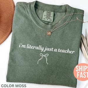 May include: A moss green t-shirt with the text "i'm literally just a teacher" in white. A white bow graphic is below the text. A gold necklace with a pendant is on the shirt. The shirt is labeled "Comfort Colors".
