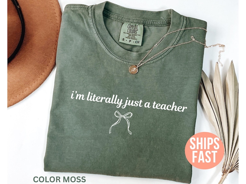 May include: A moss green t-shirt with the text "i'm literally just a teacher" in white. A white bow graphic is below the text. A gold necklace with a pendant is on the shirt. The shirt is labeled "Comfort Colors".