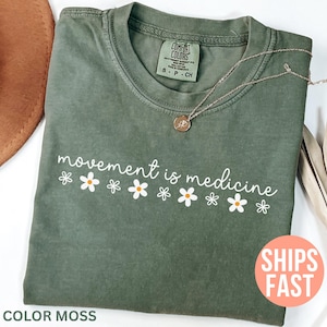 May include: A moss green t-shirt with the phrase "movement is medicine" in white script, decorated with white and yellow daisy graphics. A gold necklace and a brown hat are also visible.
