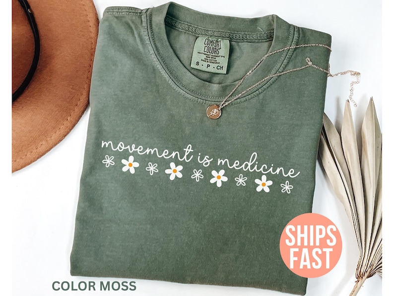 May include: A moss green t-shirt with the phrase "movement is medicine" in white script, decorated with white and yellow daisy graphics. A gold necklace and a brown hat are also visible.