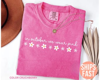 May include: A pink t-shirt with the text "in october we wear pink" and daisy graphics. A gold necklace and a brown hat are also in the image. The shirt is a Comfort Colors brand.