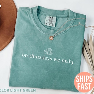 以下が含まれることがあります： 「on thursdays we mahj」の白文字が入ったライトグリーンのTシャツ。 金色のペンダント付きネックレスがシャツの上にあります。 シャツは折りたたまれ、茶色の帽子と乾燥した植物と一緒に表示されています。