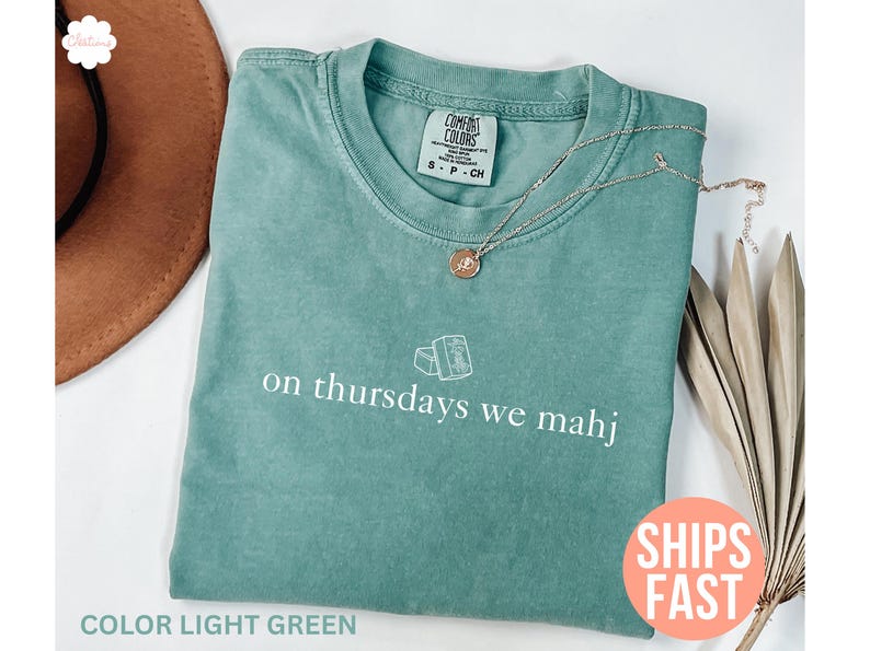以下が含まれることがあります： 「on thursdays we mahj」の白文字が入ったライトグリーンのTシャツ。 金色のペンダント付きネックレスがシャツの上にあります。 シャツは折りたたまれ、茶色の帽子と乾燥した植物と一緒に表示されています。