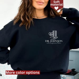 May include: Black sweatshirt with a white graphic of a spine, heart, and hands, with the text "DR. JOHNSON chiropractor". The sweatshirt has a crew neck and long sleeves. The words "Soft material" and "More color options" are also visible.