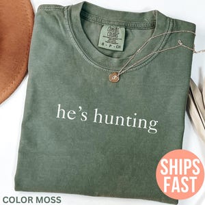 May include: A moss green t-shirt with the phrase "he's hunting" printed in white. A gold-colored necklace with a pendant rests on the shirt. A brown hat and dried plant are in the background.