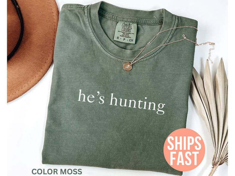 May include: A moss green t-shirt with the phrase "he's hunting" printed in white. A gold-colored necklace with a pendant rests on the shirt. A brown hat and dried plant are in the background.