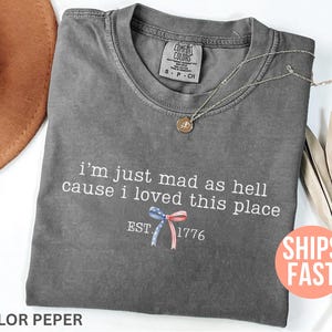 以下が含まれることがあります： 「I'm just mad as hell cause I loved this place」のテキストとリボンのグラフィックがプリントされたグレーのComfort Colors Tシャツ。ゴールドのペンダントネックレスと茶色の帽子も写っています。