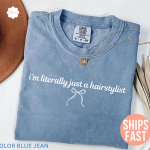 May include: A blue jean coloured t-shirt with the text "i'm literally just a hairstylist" in white. A gold necklace rests on the shirt. The shirt is folded and placed on a white surface. A brown hat is visible in the background.
