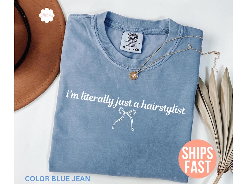 May include: A blue jean coloured t-shirt with the text "i'm literally just a hairstylist" in white. A gold necklace rests on the shirt. The shirt is folded and placed on a white surface. A brown hat is visible in the background.