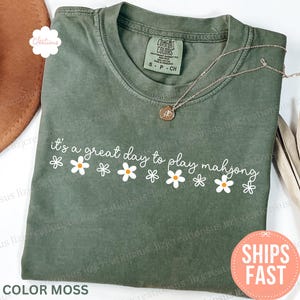 May include: A moss green t-shirt with the text "it's a great day to play mahjong" in white script and daisy graphics. A gold necklace with a pendant is draped on the shirt. The shirt is labeled "Comfort Colors".