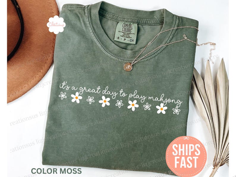 May include: A moss green t-shirt with the text "it's a great day to play mahjong" in white script and daisy graphics. A gold necklace with a pendant is draped on the shirt. The shirt is labeled "Comfort Colors".
