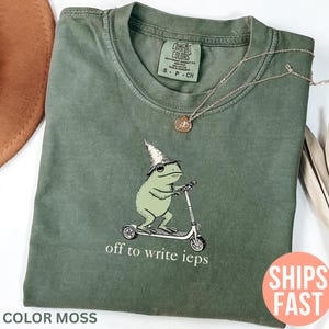 Könnte beinhalten: Grünes Comfort Colors T-Shirt mit einer Illustration einer Froschfigur mit Hexenhut auf einem Roller. Der Text "off to write ieps" ist darunter gedruckt. Die Farbe des Shirts ist Moosgrün.